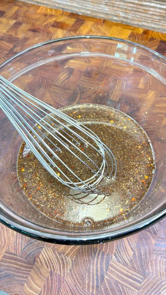 A metal whisk sits in a glass bowl containing a mixture of seasonings and liquid, with visible spices and pepper flakes, on a patterned wooden surface.