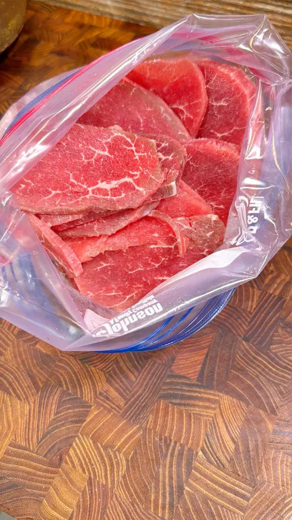 Raw slices of red beef placed inside a resealable plastic bag, resting on a wooden countertop with a distinctive geometric pattern.