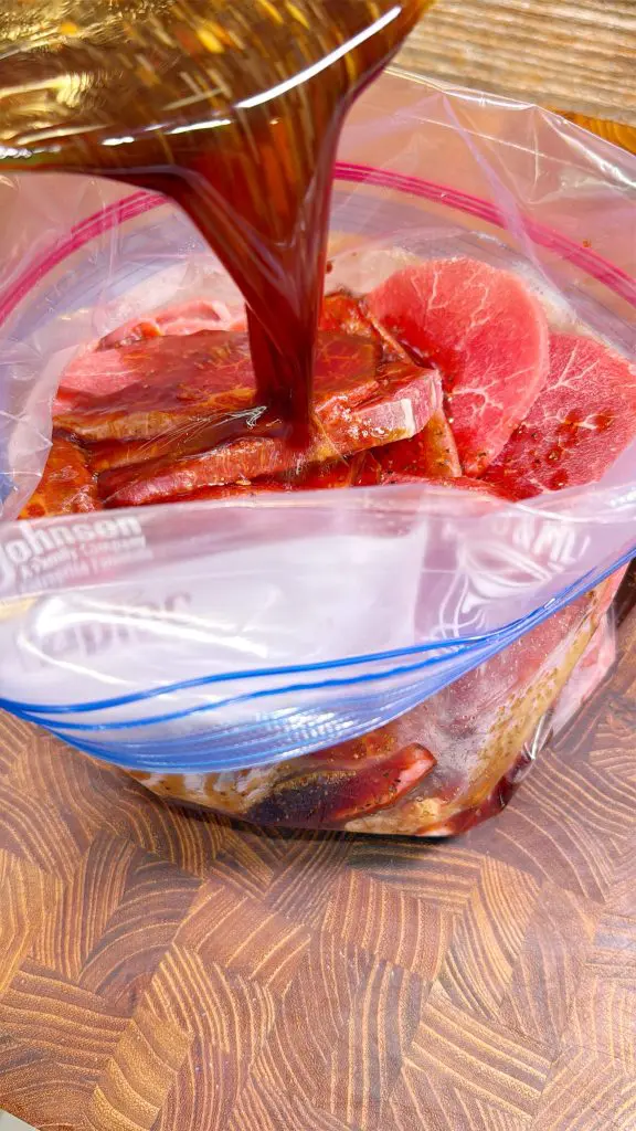 Marinade for dr pepper beef jerky being poured over raw steak slices in a clear plastic zip-top bag, which is placed on a wooden cutting board.