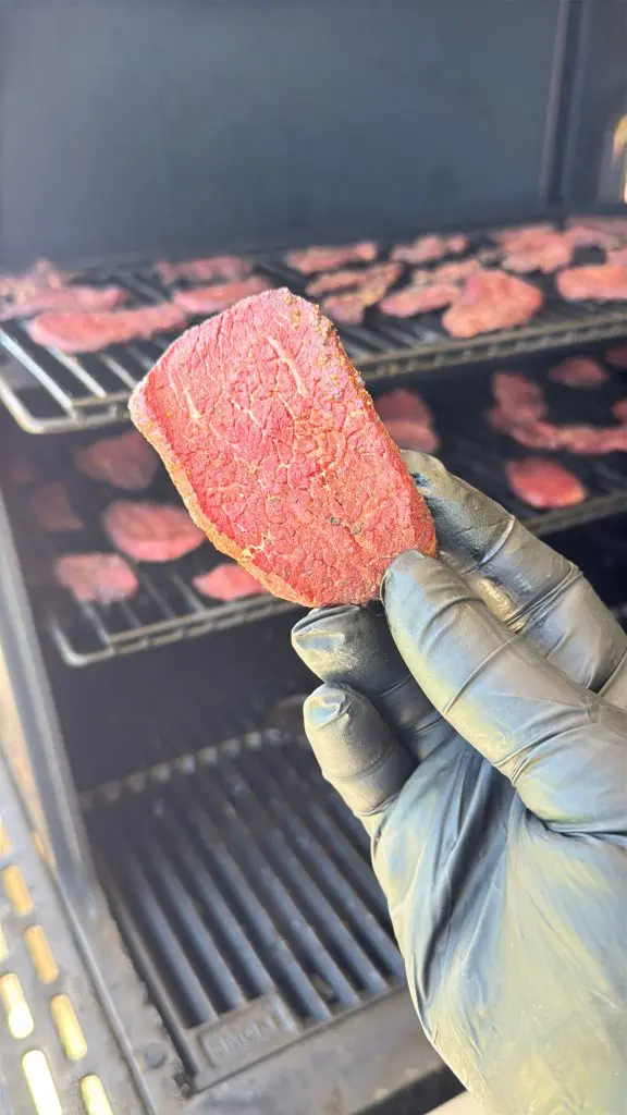 A gloved hand holds a thin slice of raw Dr Pepper beef jerky in front of a smoker filled with additional slices drying on metal racks.