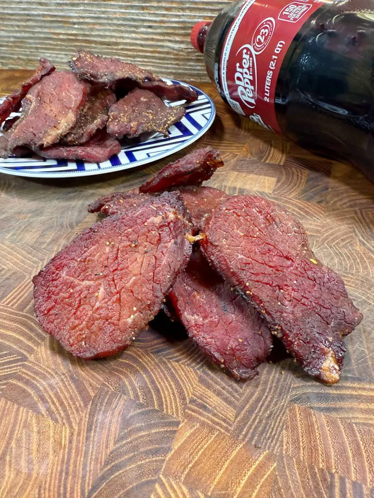 Close-up of slices of beef jerky on a wooden surface, with a plate of more jerky in the background and a bottle of Dr Pepper soda lying on its side. Recipe is for Dr. Pepper Beef Jerky