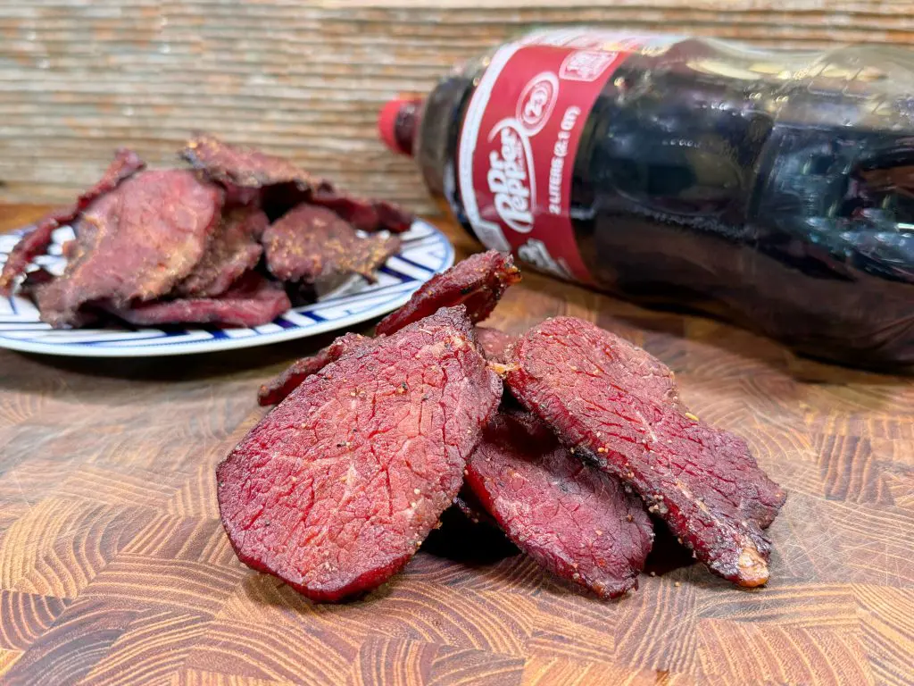 Sliced pieces of red beef jerky are arranged on a wooden surface, with more jerky on a plate in the background. A bottle of Dr Pepper soda lies on its side behind the jerky.