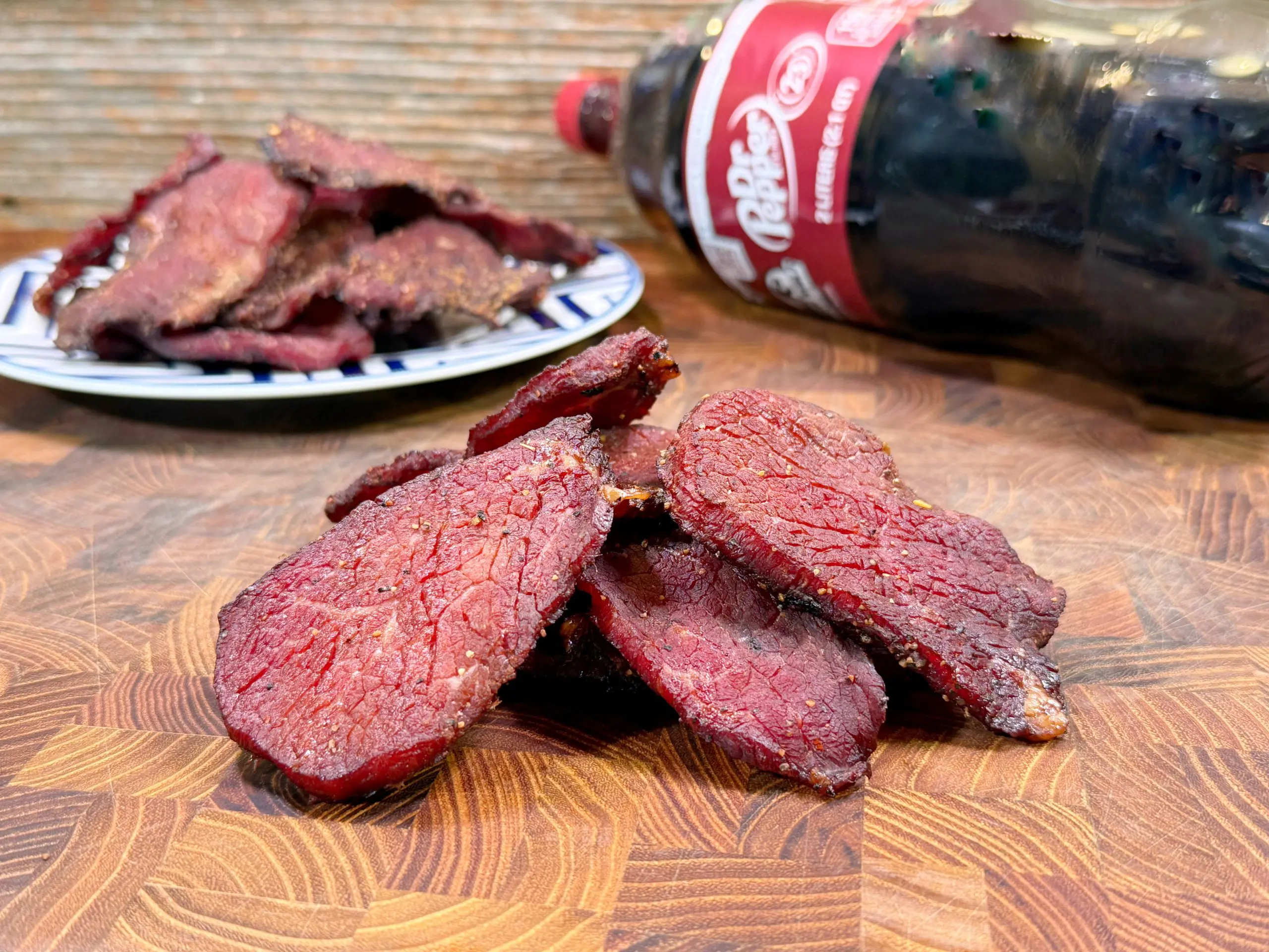 Slices of beef jerky stacked on a wooden surface with a plate of more jerky and a bottle of Dr Pepper soda in the background.