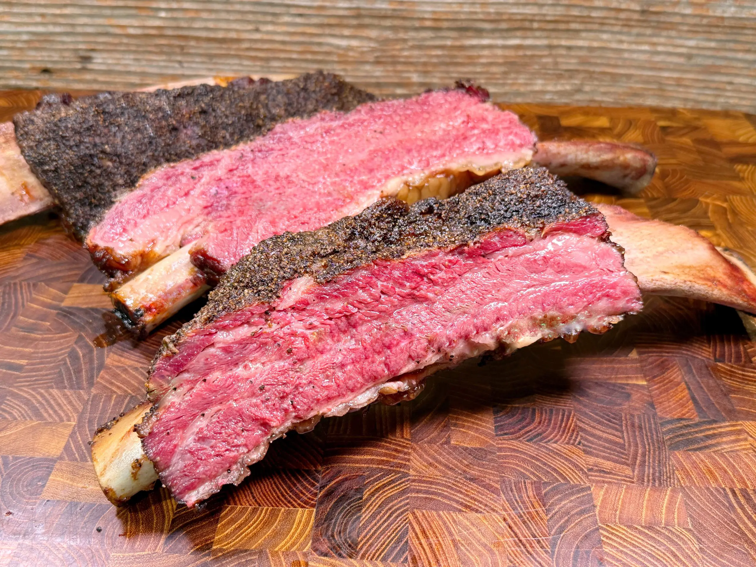 Two large, juicy beef ribs with a pink, medium-rare center and a dark, seasoned crust rest on a wooden cutting board.