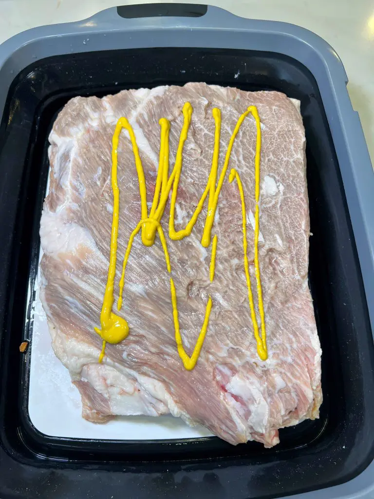A large, raw slab of pork belly in a black tray, topped with several zigzag lines of yellow mustard, ready to be smoked like pastrami beef ribs.
