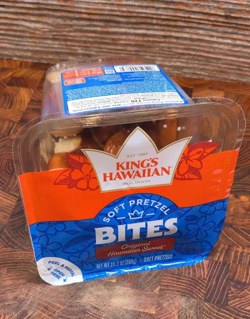 A clear plastic container of King’s Hawaiian Stuffed Pretzel Bites, featuring red, blue, and orange packaging with floral designs, sits on a wooden surface. The stuffed pretzel bites are visible inside the container.