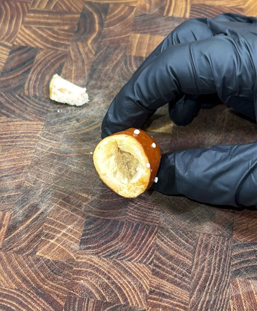 A hand in a black glove holds a small, partially hollowed pretzel bite with a piece missing. The pretzel rests on a wooden, patterned surface with another small piece nearby.