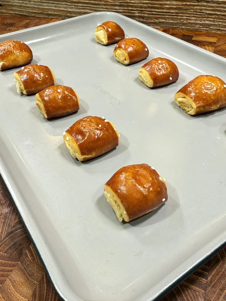 Nine small pretzel-wrapped snacks with a golden-brown crust are spaced out on a light gray baking sheet, resting on a wooden surface. Some snacks are sprinkled with coarse salt.