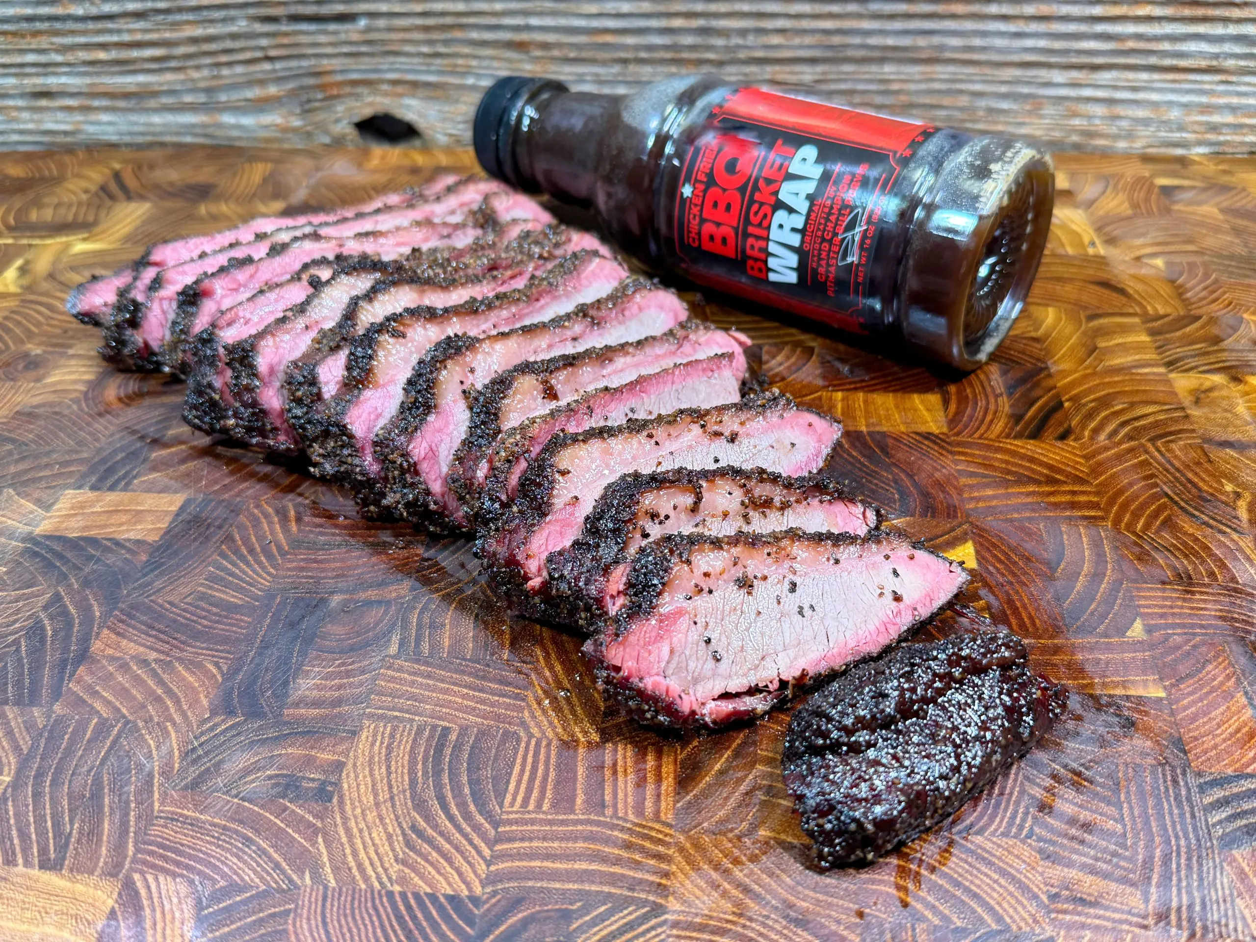 Sliced, juicy smoked meat with a peppery crust is arranged on a wooden cutting board. A bottle labeled “BBQ Wrap” lies in the background, partially on its side.