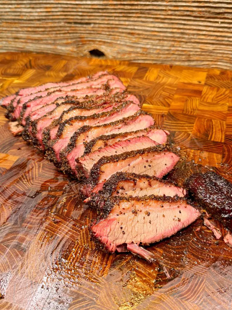 Sliced smoked brisket with a dark peppery crust arranged on a wooden cutting board, showing a juicy interior and visible smoke ring.