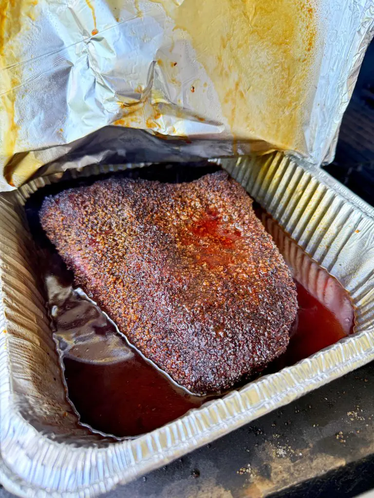 A cooked, seasoned brisket sits in a disposable aluminum tray filled with juices, partially covered by crumpled foil. The brisket has a dark, flavorful crust from the rub.