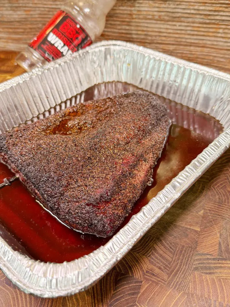 A seasoned, cooked brisket sits in a disposable aluminum tray with juices at the bottom. An empty plastic BBQ seasoning bottle lies in the background on a wooden surface.
