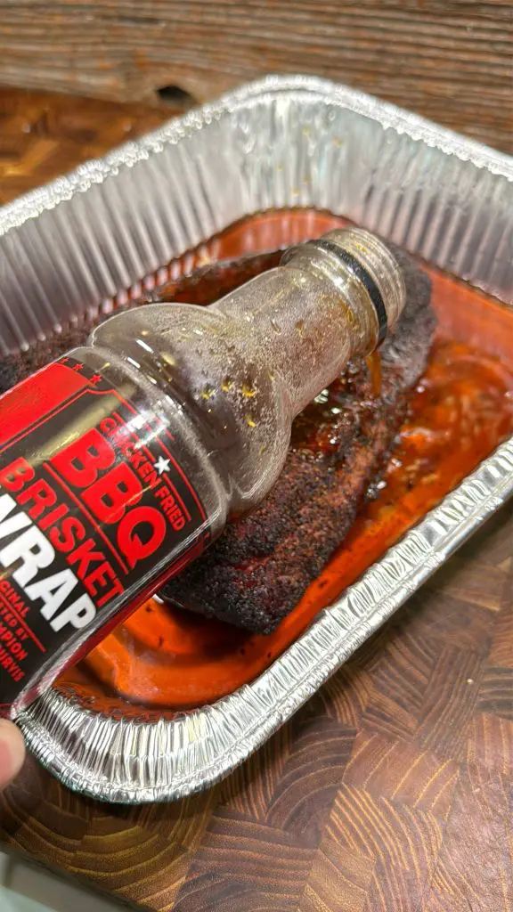 A hand pours BBQ Brisket Wrap sauce from a bottle onto a cooked brisket in a foil tray, with sauce pooling around the meat on a wooden surface.