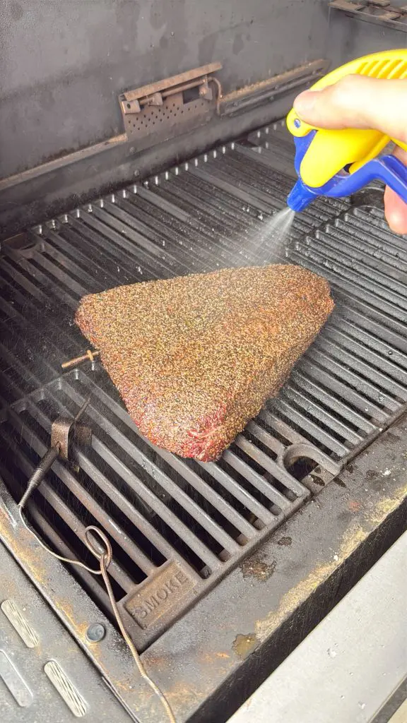 A seasoned brisket is being sprayed with liquid from a yellow and blue spray bottle while cooking on a barbecue grill.