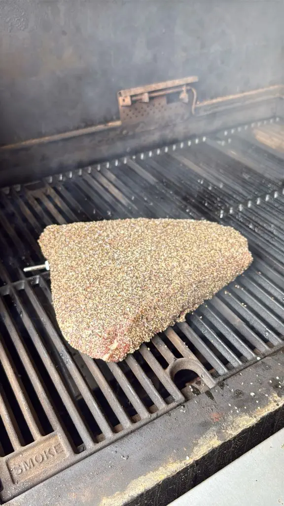 A seasoned brisket cooks on the grates of a smoker grill, surrounded by rising smoke.