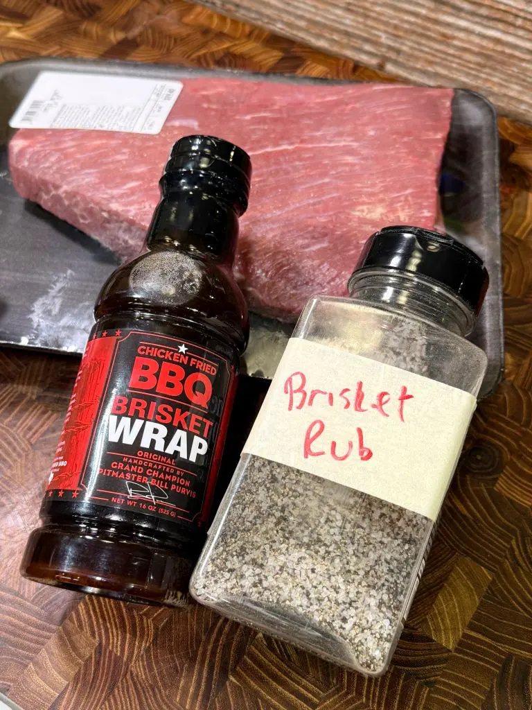 A raw brisket on a tray sits behind a bottle of BBQ brisket wrap sauce and a jar labeled Brisket Rub on a wooden cutting board.