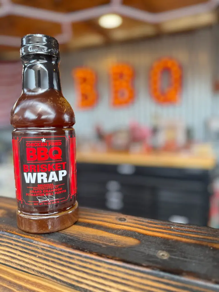 A bottle of Chicken Fried Brisket Wrap BBQ sauce sits on a wooden counter, with a blurred background featuring large orange BBQ letters and a rustic interior.
