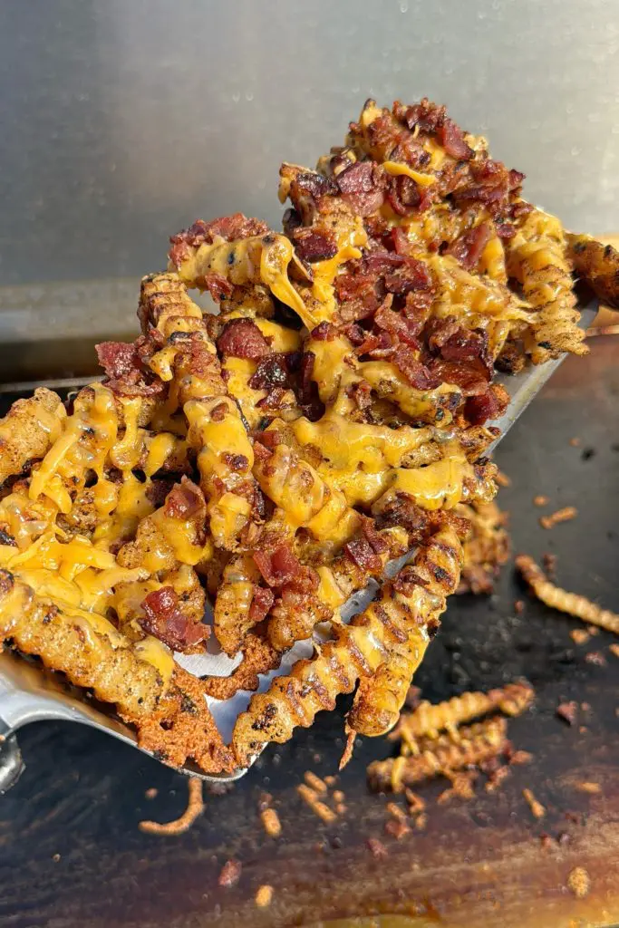 A close-up of bacon fries with crinkle-cut potatoes, smothered in melted cheddar cheese and crispy bacon bits, served on a metal spatula.
