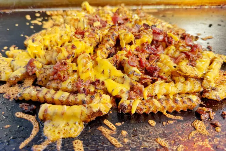 Crinkle-cut Bacon French Fries topped with melted cheese and crispy bacon pieces, cooked to perfection on a hot griddle for the ultimate Griddle Recipe, with cheese and savory seasoning scattered around.