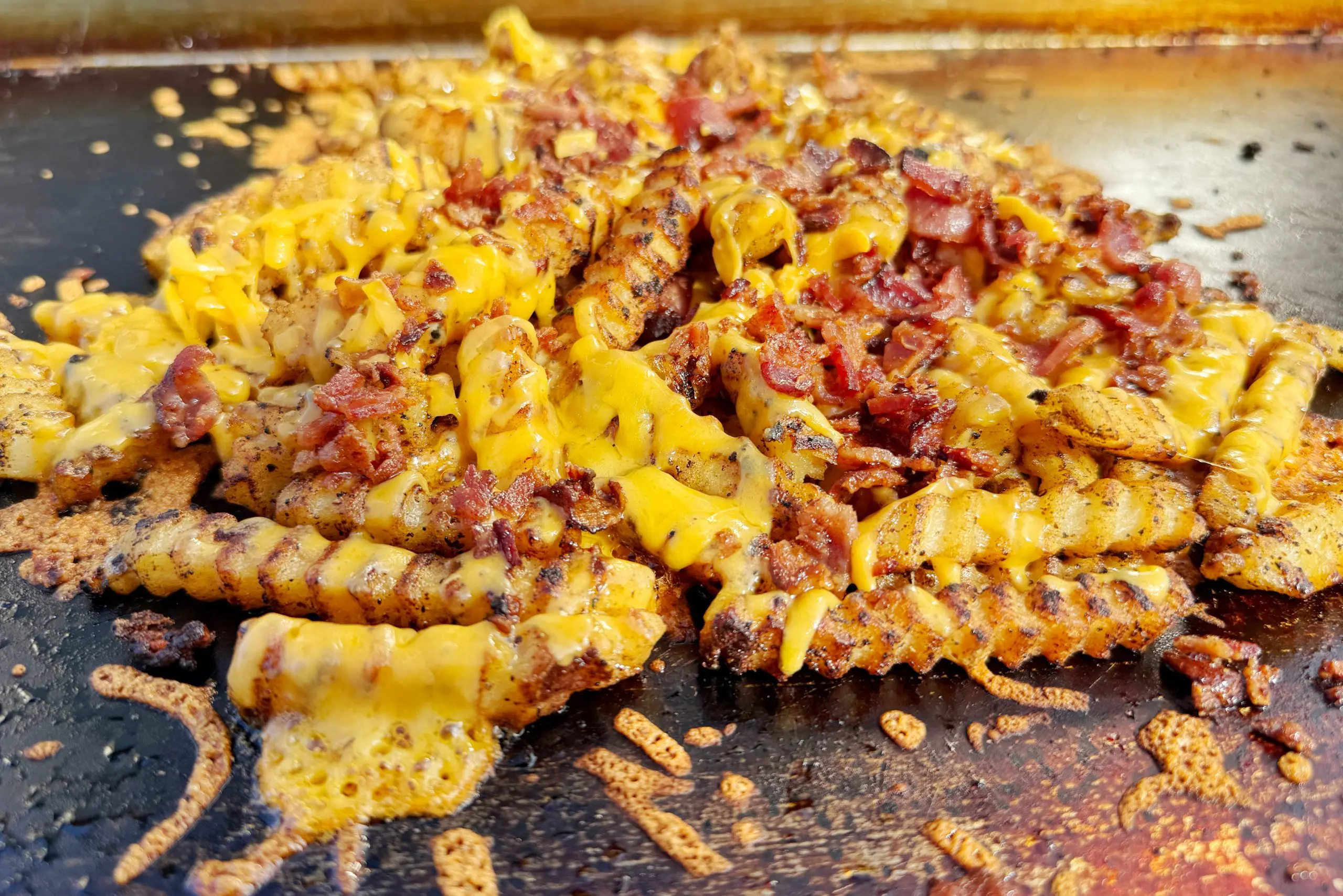 Crinkle-cut Bacon French Fries topped with melted cheese and crispy bacon pieces, cooked to perfection on a hot griddle for the ultimate Griddle Recipe, with cheese and savory seasoning scattered around.