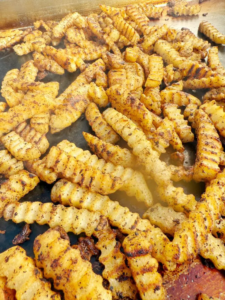 Crinkle-cut French fries with golden, crispy edges and visible seasoning are spread out on a flat cooking surface, topped with melted cheese and savory bacon for a warm, irresistibly appetizing appearance.