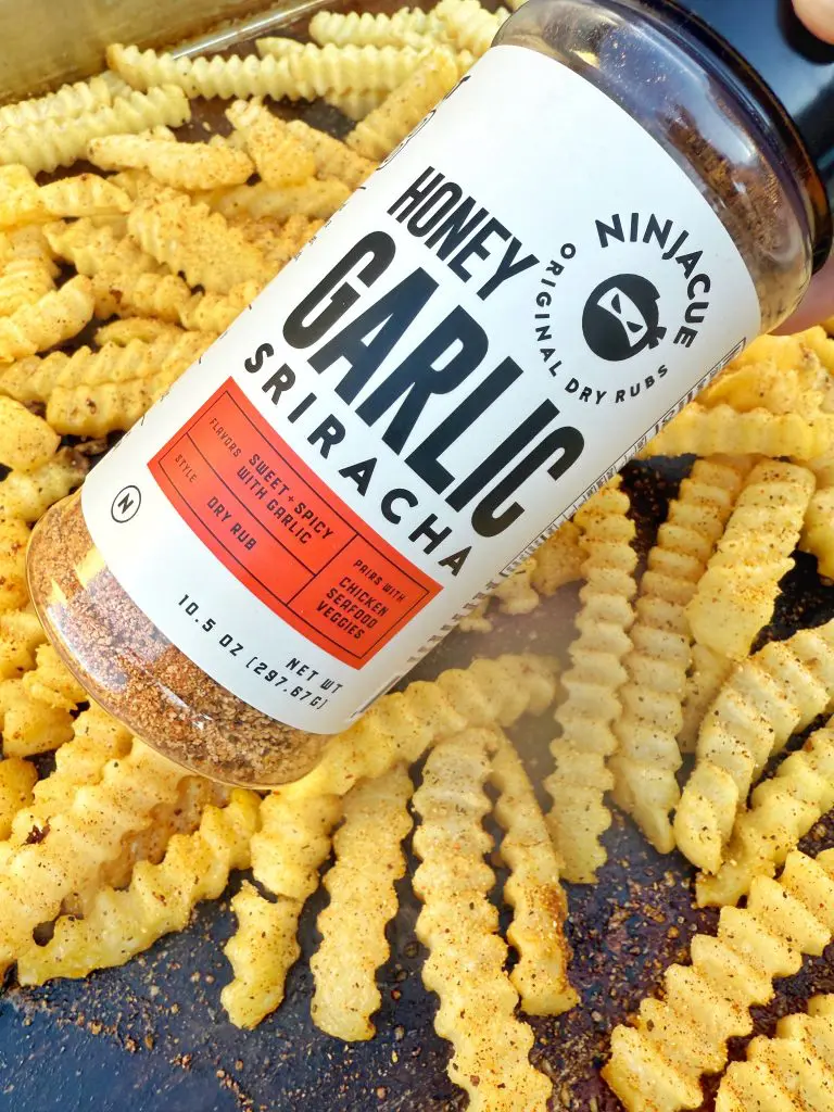 A container of Ninjacue Honey Garlic Sriracha dry rub is held above crinkle-cut fries, perfect for a bold griddle recipe or transforming into loaded cheese fries, all seasoned and spread out on a sizzling cooking surface.