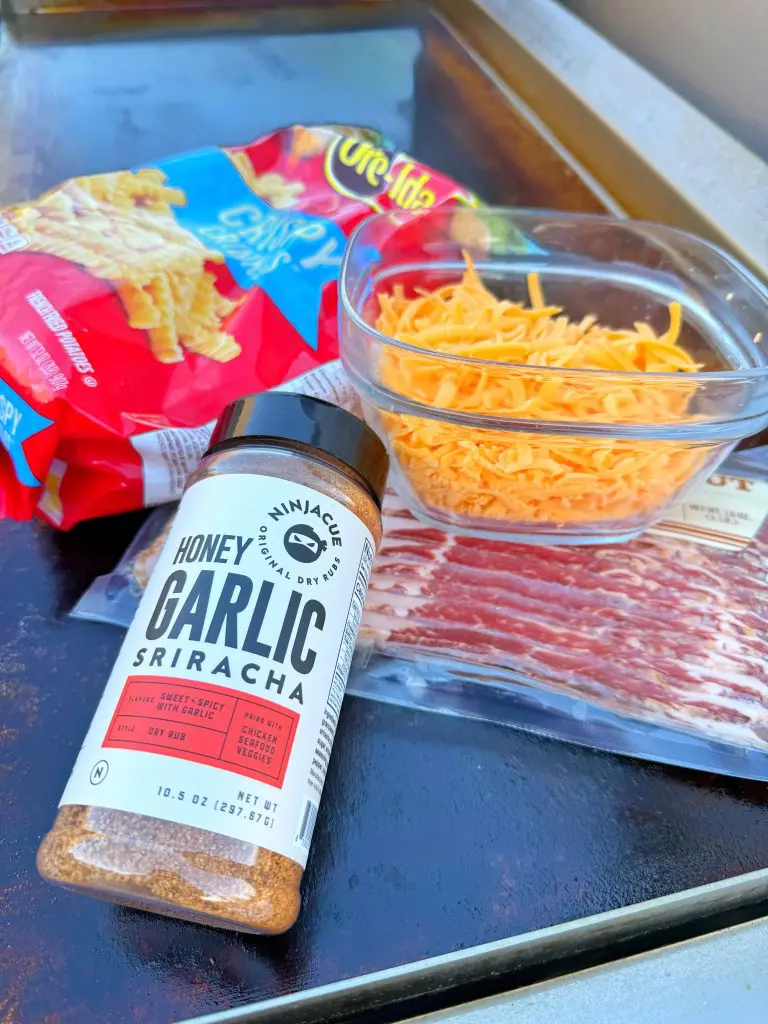 A bottle of honey garlic sriracha seasoning, a bag of crinkle-cut fries, shredded cheddar cheese, and uncooked bacon are arranged on a flat cooking surface—perfect for a bacon fries griddle recipe.
