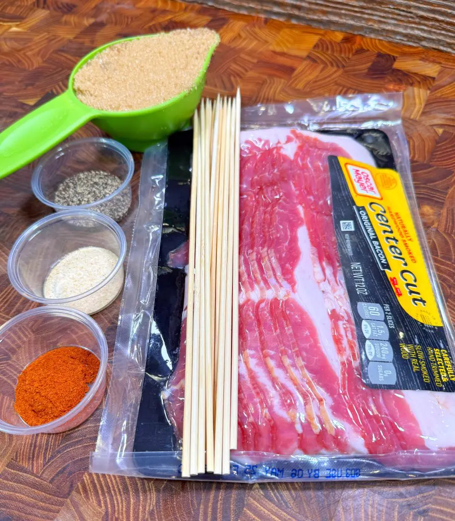 A package of center cut bacon, wooden skewers, a green scoop of brown sugar, and small containers of paprika, garlic powder, and pepper are arranged on a wooden surface.