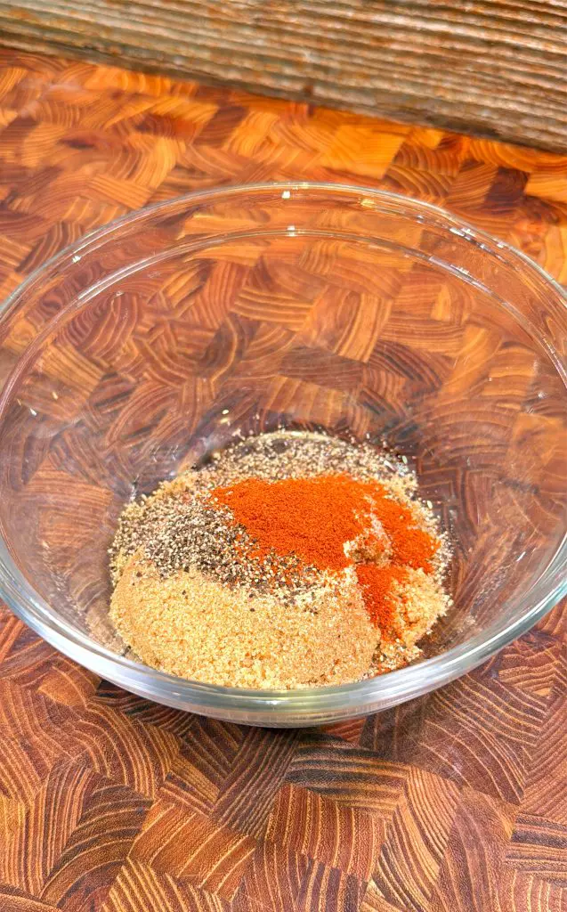 A glass bowl on a wooden surface contains a mixture of assorted spices, including black pepper, paprika, and granulated seasonings in separate piles.