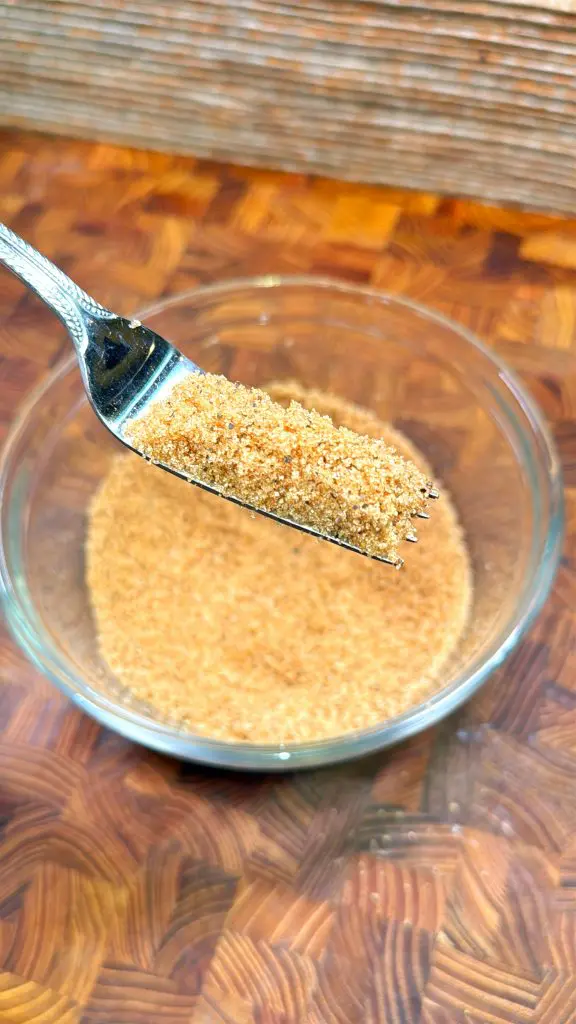 A close-up of a fork holding a small amount of brown sugar above a glass bowl filled with more brown sugar, set on a wooden surface.