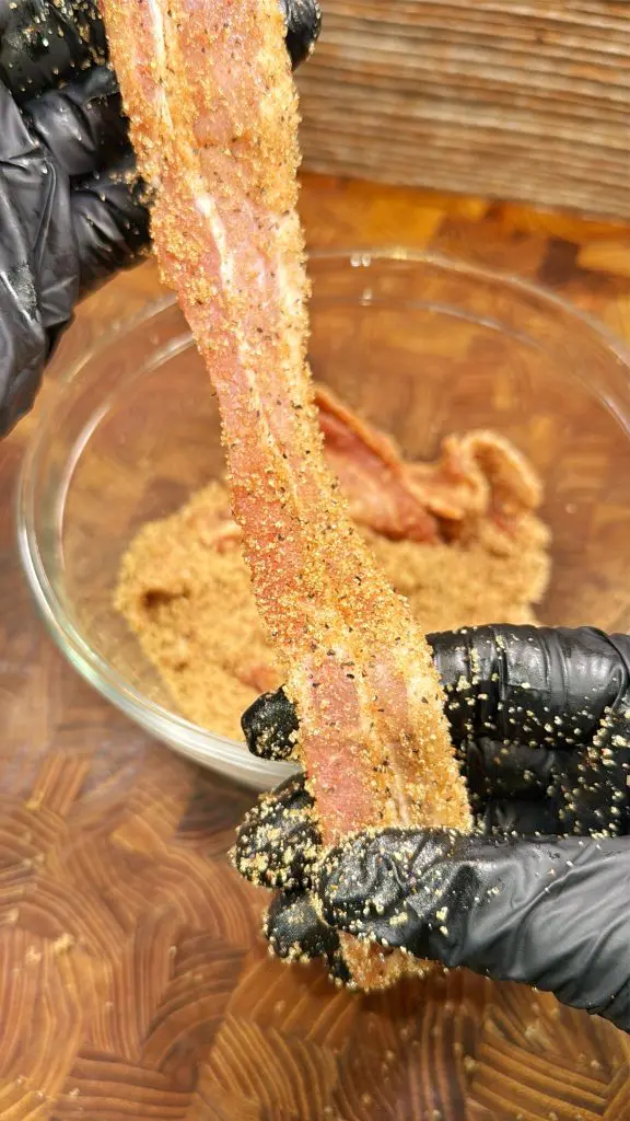 A person wearing black gloves coats a strip of raw bacon with a mixture of spices and sugar over a glass bowl on a wooden surface.
