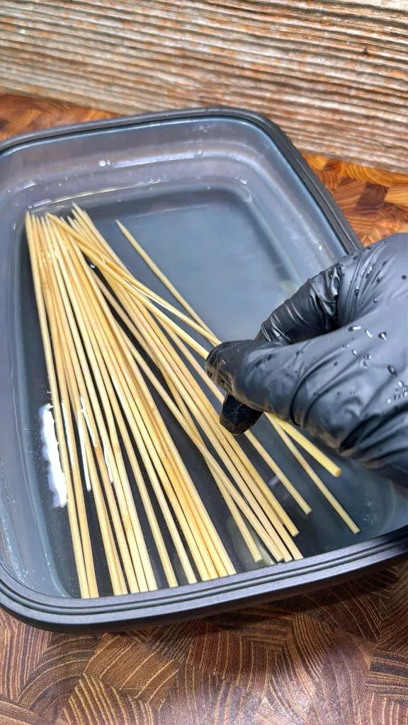 A hand wearing a black glove is placing wooden skewers into a container filled with water to soak them. The container rests on a wooden surface.