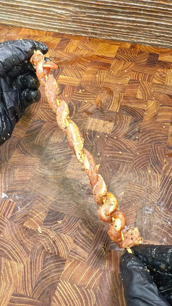 A person wearing black gloves holds a skewer with pieces of seasoned, twisted meat over a wooden cutting board. These are bacon twists.
