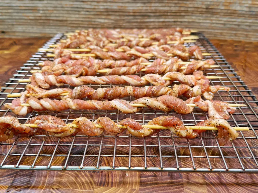 Rows of marinated, twisted strips of meat are threaded onto skewers and arranged on a metal cooling rack, ready to be grilled, with a rustic wooden background beneath.