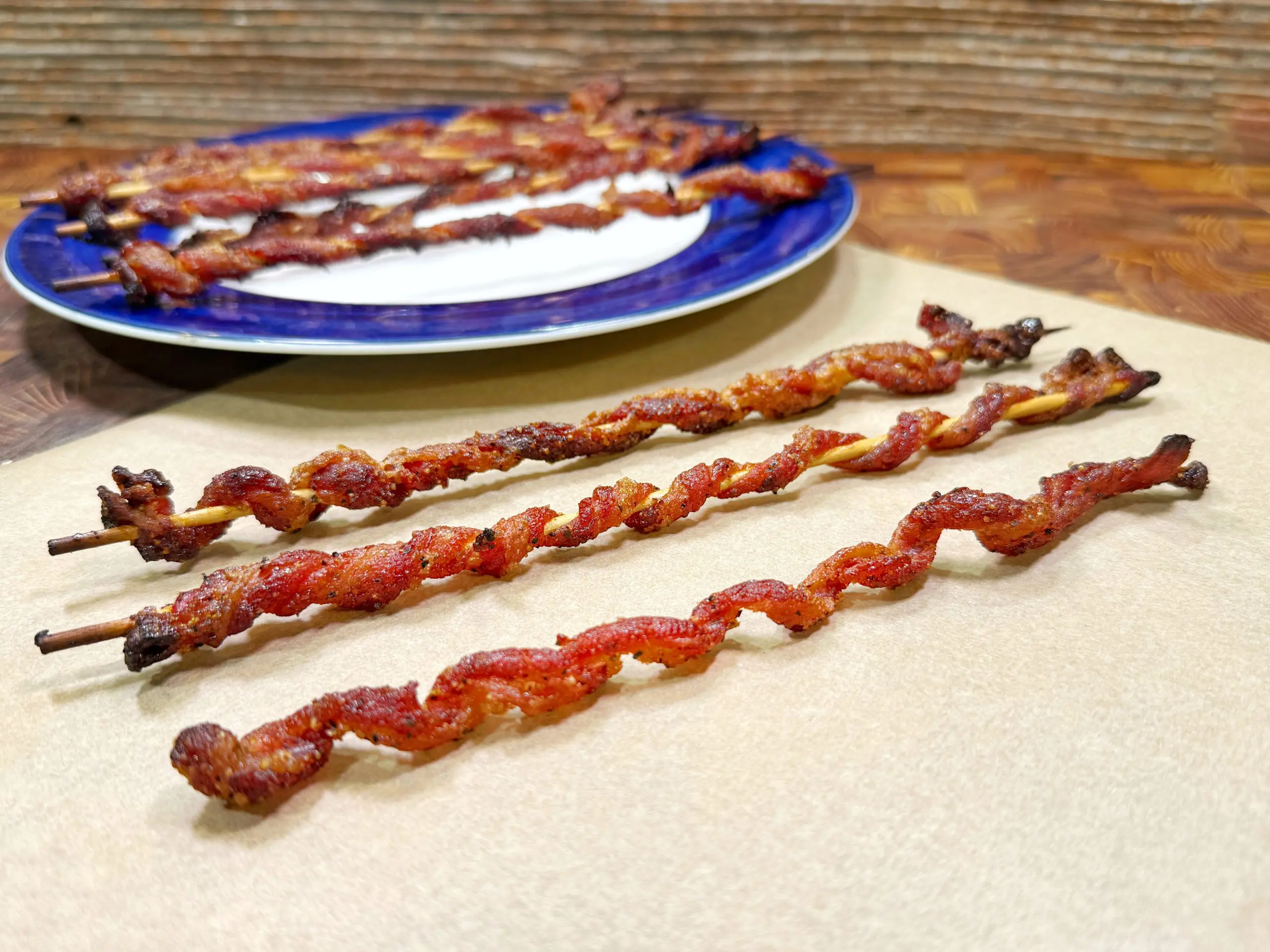 Four twisted strips of cooked bacon are skewered on sticks and laid out on parchment paper, with more bacon skewers on a blue-rimmed plate in the background. The surface and backdrop are wooden.