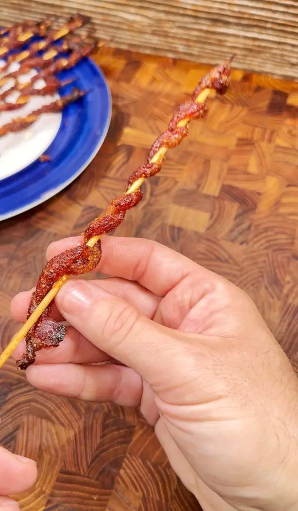 A hand holds a wooden skewer with a spiral strip of cooked bacon wrapped around it. In the background, a blue plate with more bacon skewers sits on a wooden cutting board.