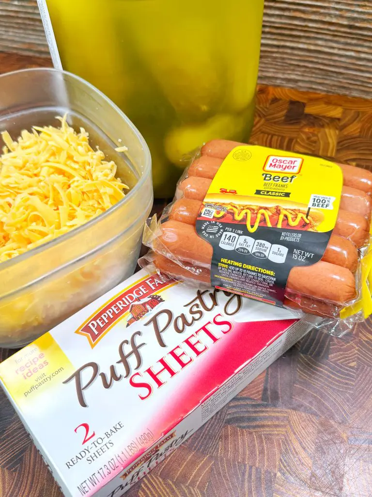 A package of Oscar Mayer beef franks, a box of Pepperidge Farm puff pastry sheets, a bowl of shredded cheese, and a large jar of pickles on a wooden surface.