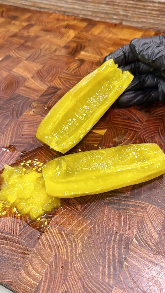 A gloved hand holds a halved pickle on a wooden cutting board, with seeds and pulp scooped out and placed next to it.
