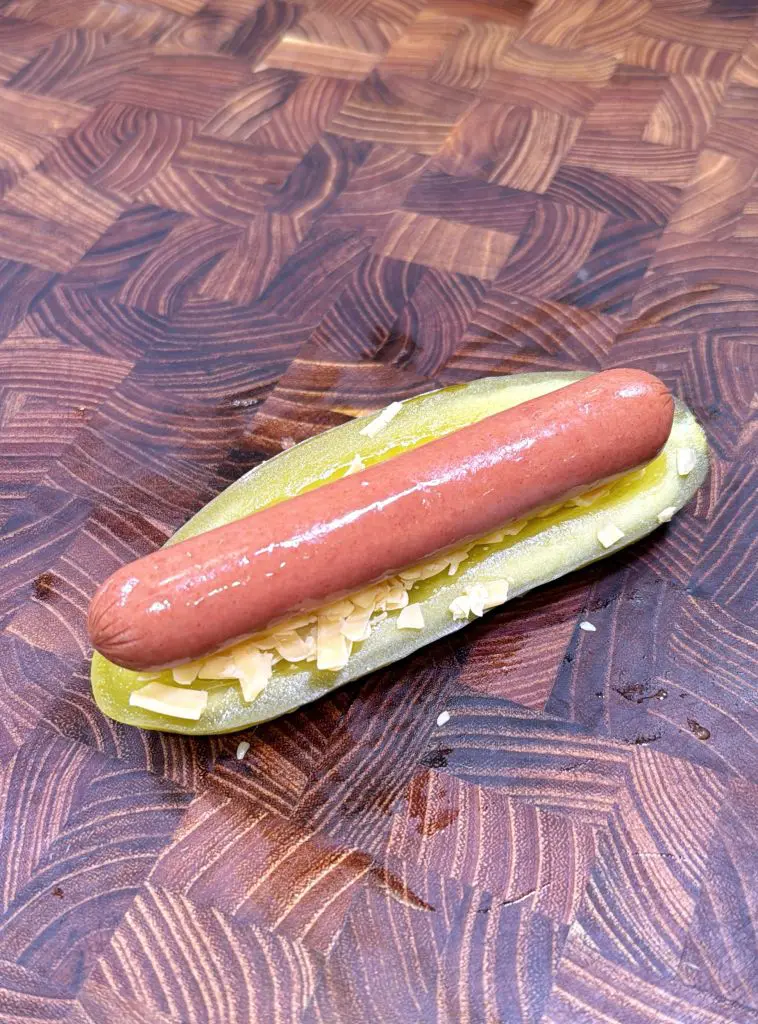 A hot dog sausage sits on shredded cheese inside a sliced pickle, replacing a traditional bun, on a patterned wooden surface. Making a pickles dogs recipe