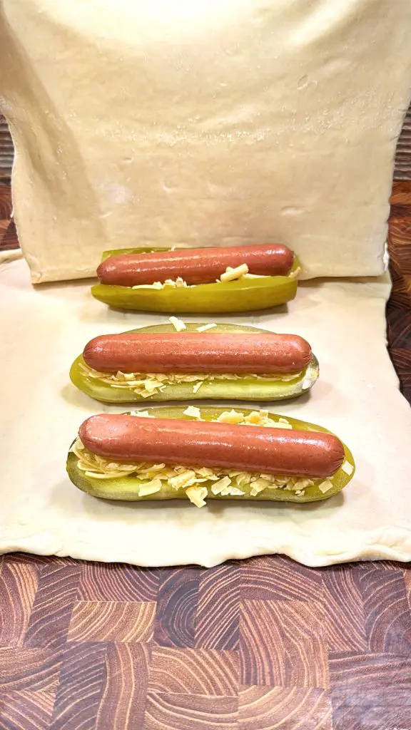 Three hot dogs placed inside halved pickles, topped with shredded cheese, are arranged on a sheet of uncooked dough on a wooden surface.