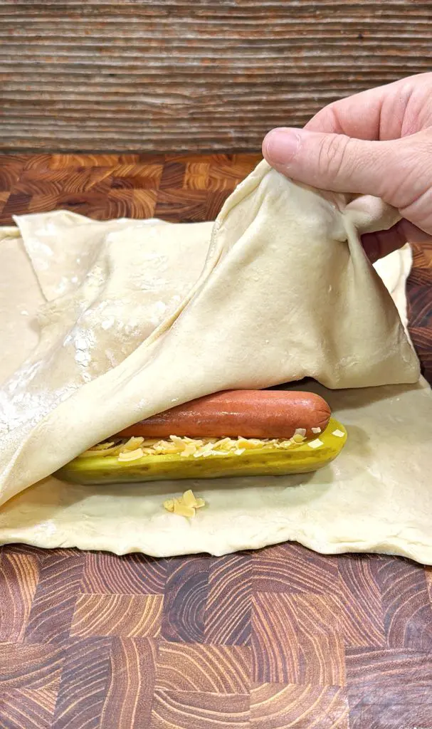 A hand lifts a sheet of dough over a pickle stuffed with shredded cheese and a hot dog, all placed on a wooden surface, preparing to wrap the ingredients with the dough.