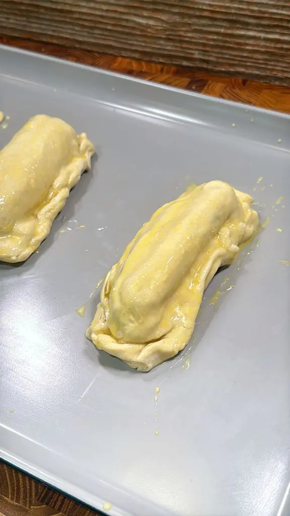 Two unbaked pastry-wrapped items coated with egg wash rest on a gray baking sheet, ready to be baked. The pastries have a rustic, homemade appearance with slightly uneven edges.