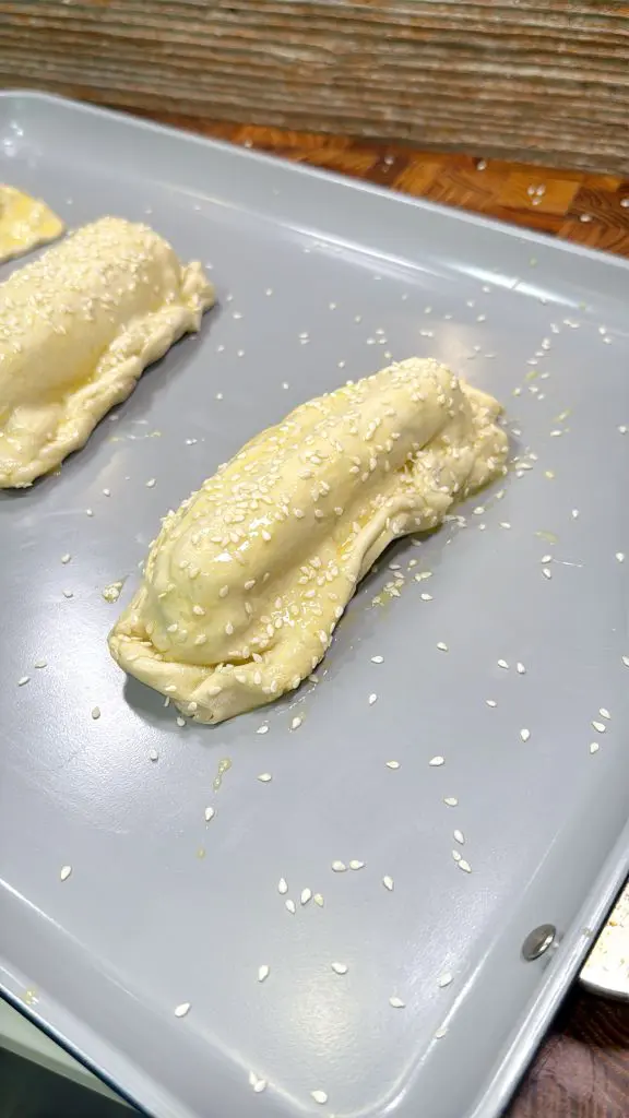 Unbaked pastry filled with a stuffing, topped with sesame seeds, placed on a gray baking tray, ready to be baked.