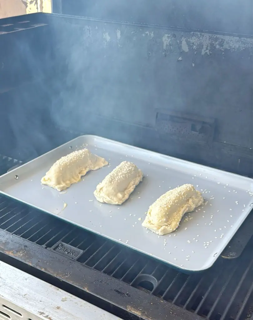 Three unbaked, sesame seed-topped pastries are on a light-colored baking sheet inside a smoky grill or oven. The pastries are spaced apart, and the surrounding area appears to be a BBQ grill.