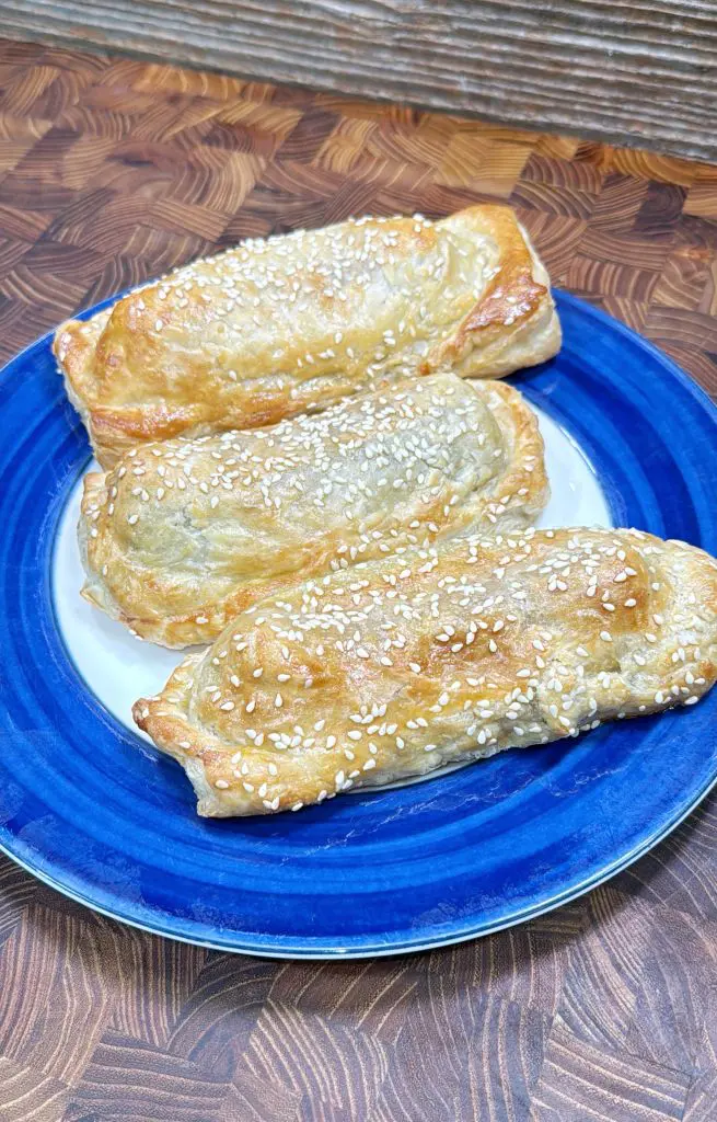 Three golden-brown, flaky pastries topped with sesame seeds are arranged on a blue and white plate, sitting on a wooden surface.
