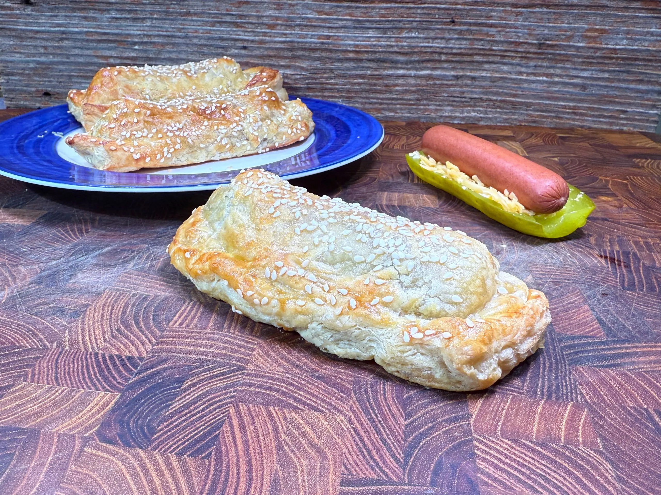 A flaky pastry topped with sesame seeds sits on a wooden surface in the foreground. Behind it, a blue plate holds more pastries, and a hot dog rests on a sliced pickle. The background is rustic wood.