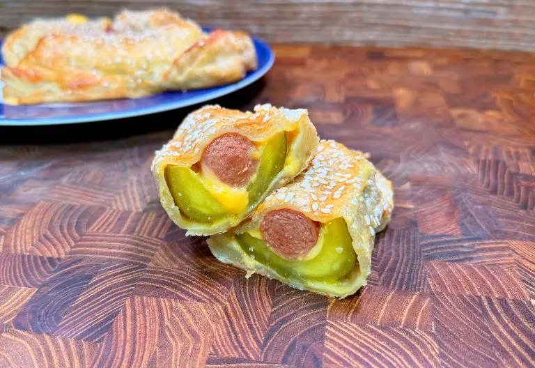 A close-up of two pastry-wrapped hot dogs with visible pickles and melted cheese inside, topped with sesame seeds, stacked on a wooden surface. A whole pastry is blurred in the background on a blue plate.