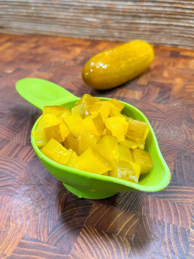 A green measuring spoon filled with diced yellow pickles sits on a wooden surface, with a whole pickle in the background.