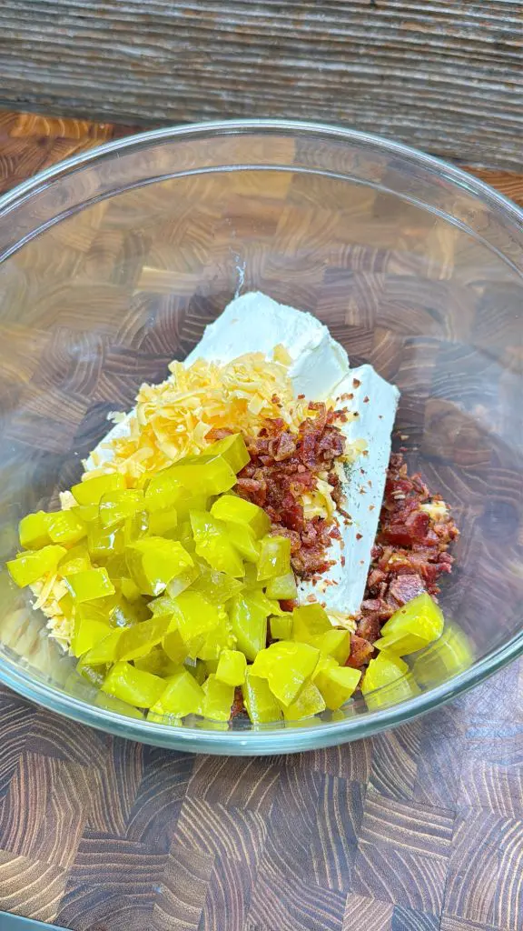 A glass bowl on a wooden surface containing cream cheese, shredded cheese, chopped bacon, and diced pickles, ready to be mixed together.