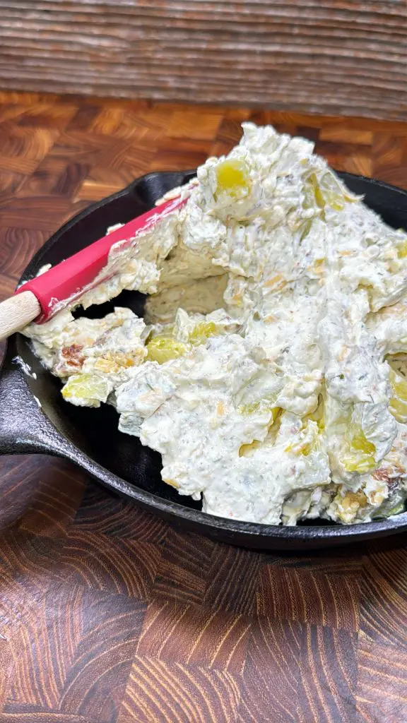 A black skillet with a creamy mixture containing chopped green pickles and visible seasoning, being mixed with a red spatula, sits on a dark wooden surface.